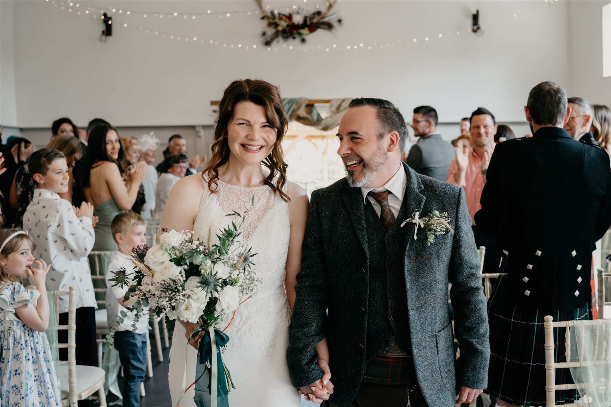 A bride and groom walk down the aisle in Netherdale House after their wedding. They smile for a photograph.